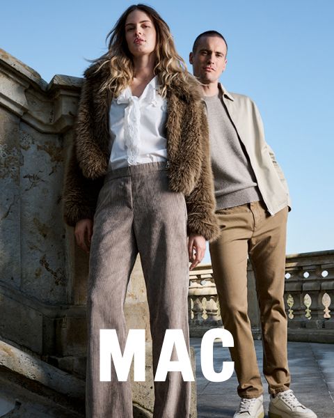 Eine Frau in einer Pelzjacke und weiten Hosen steht neben einem Mann in einer beigen Jacke und khakifarbenen Hosen. Beide posieren selbstbewusst im Freien vor einem steinernen Geländer unter blauem Himmel. Das Wort "MAC" erscheint am unteren Rand.