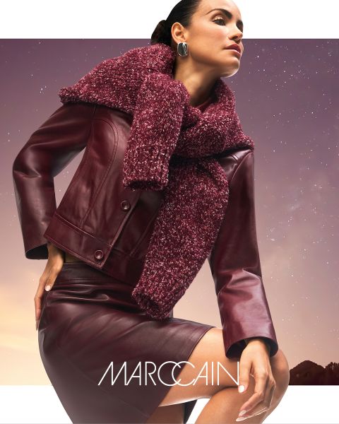 Eine Frau posiert selbstbewusst in einer weinroten Lederjacke und einem Rock mit einem klobigen, strukturierten weinroten Schal. Im Hintergrund ist ein düsterer Himmel zu sehen, und der Markenname "Marc Cain" ist am unteren Rand zu sehen.