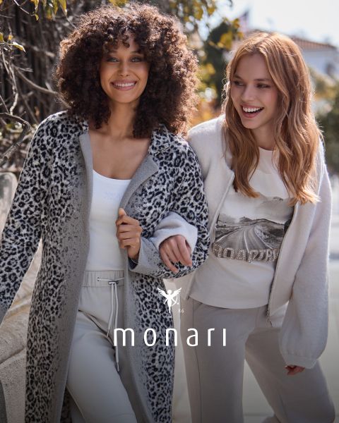 Zwei lächelnde Frauen gehen im Freien spazieren. Beide tragen helle, modische Monari-Kleidung. Die eine trägt einen Mantel mit Leopardenmuster, die andere ein Sweatshirt mit Grafik und eine Strickjacke. Unten ist das Monari-Logo zu sehen.