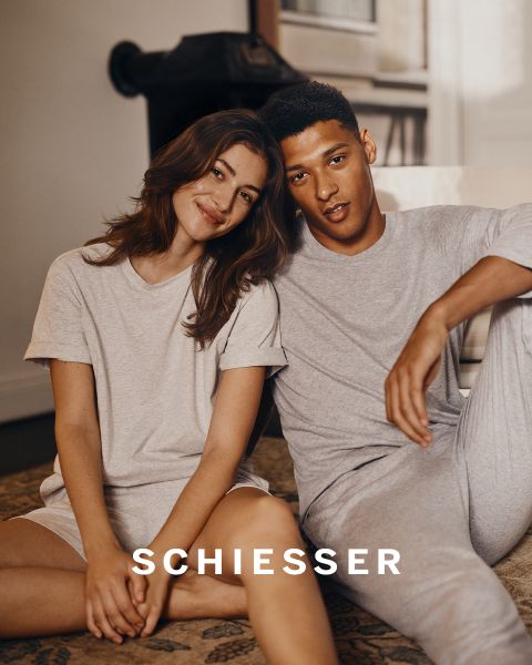 Eine Frau und ein Mann in hellgrauer Loungewear sitzen dicht nebeneinander auf einem Teppich und lächeln sanft in die Kamera. Der Markenname "SCHIESSER" ist im unteren Teil des Bildes zu sehen.