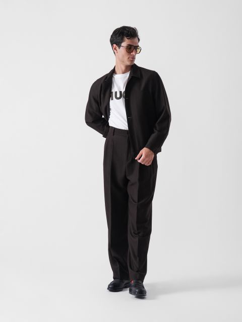 Ein Mann, der eine Sonnenbrille, ein weißes T-Shirt mit Text, eine dunkle Jacke und eine dazu passende dunkle Hose trägt, steht vor einem schlichten weißen Hintergrund und blickt mit einer Hand auf dem Rücken nach links.
