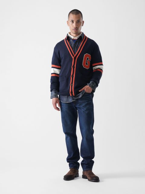 Ein Mann steht mit dem Gesicht nach vorne und trägt eine marineblaue Strickjacke mit orangefarbenen Streifen und dem großen Buchstaben "G" über einem Jeanshemd und Jeans, mit braunen Stiefeln, vor einem schlichten weißen Hintergrund.