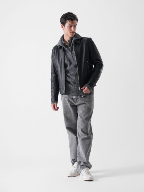Ein Mann steht vor einem schlichten weißen Hintergrund, trägt eine schwarze Lederjacke über einem grauen Kapuzenpullover und Hemd, graue Jeans und weiße Turnschuhe und blickt mit einer Hand in der Tasche nach links.