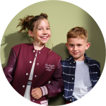 Zwei lächelnde Kinder stehen Seite an Seite vor einem grünen Hintergrund. Das Mädchen trägt eine kastanienbraune College-Jacke, der Junge ein marineblau kariertes Hemd über einem weißen T-Shirt. Sie sehen entspannt und fröhlich aus.