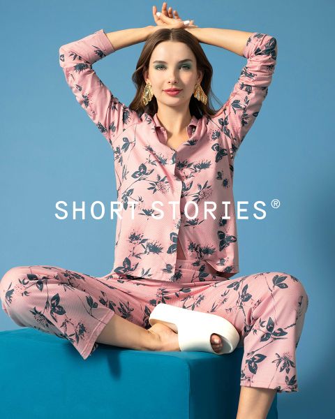 Eine Frau sitzt im Schneidersitz auf einem blauen Block, trägt ein rosa geblümtes Pyjama-Set und weiße Plateausandalen, die Arme erhoben und die Hände hinter dem Kopf. Der Text "SHORT STORIES" erscheint quer über dem Bild.