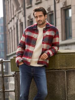 Ein Mann mit kurzen dunklen Haaren und einem Bart lehnt im Freien an einem steinernen Geländer. Er trägt eine rot-weiß karierte Jacke über einem cremefarbenen Pullover und blaue Jeans. Im Hintergrund sind städtische Gebäude zu sehen.