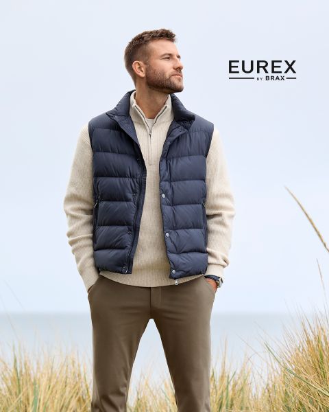 Ein Mann steht im Freien in der Nähe von hohem Gras, trägt einen beigen Pullover, eine marineblaue Pufferweste und eine braune Hose und schaut zur Seite. Das "EUREX by BRAX"-Logo erscheint auf der rechten Seite. Der Himmel ist bewölkt.