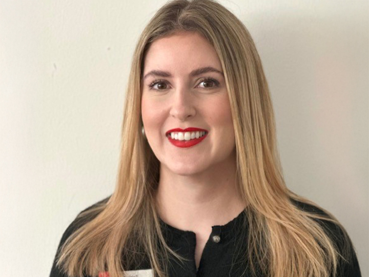 Frau mit langen blonden Haaren und rotem Lippenstift, die ein schwarzes Oberteil trägt, lächelt vor einem schlichten hellen Hintergrund.