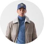 Eine Person, die eine blaue Baseballmütze, einen blauen Rollkragenpullover und eine graue Jacke mit weißem Shearling-Kragen trägt, posiert vor einem schlichten hellen Hintergrund.