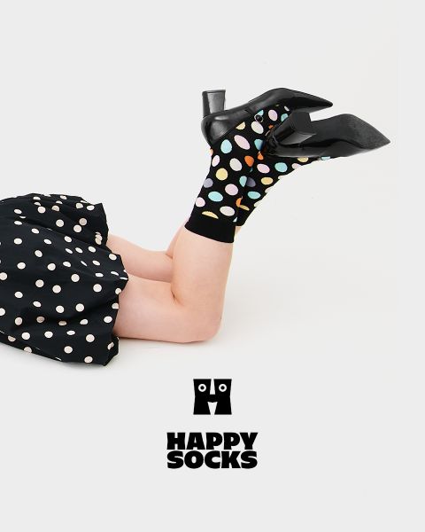 Eine Person mit schwarzen Stöckelschuhen und schwarzen Socken mit bunten Tupfen liegt auf der Seite, Beine hoch, vor einem weißen Hintergrund. Sie trägt einen schwarzen gepunkteten Rock. Darunter erscheint das Happy Socks Logo.