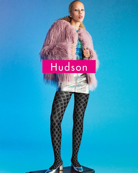 Eine Person steht auf einem Podest vor einem blauen Hintergrund und trägt einen rosa Kunstpelzmantel, ein kariertes Oberteil, einen silbernen Metallic-Rock, eine gemusterte schwarze Strumpfhose und spitze Schuhe. Das Wort "Hudson" erscheint in fetter Schrift quer über das Bild.