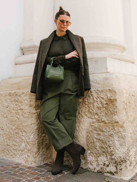 Eine Frau steht an einer Steinmauer. Sie trägt eine grüne Hose mit hoher Taille, eine dunkle Bluse, einen über die Schultern drapierten Mantel, eine Sonnenbrille, hochhackige Stiefel und eine grüne Handtasche.