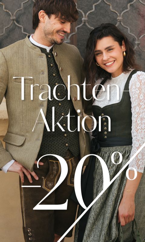 Ein Mann und eine Frau tragen traditionelle bayerische Kleidung und lächeln zusammen. Der Text auf dem Bild lautet "Trachten Aktion 20%". Im Hintergrund sind eine Steinmauer und ein dekoratives Fenster zu sehen.
