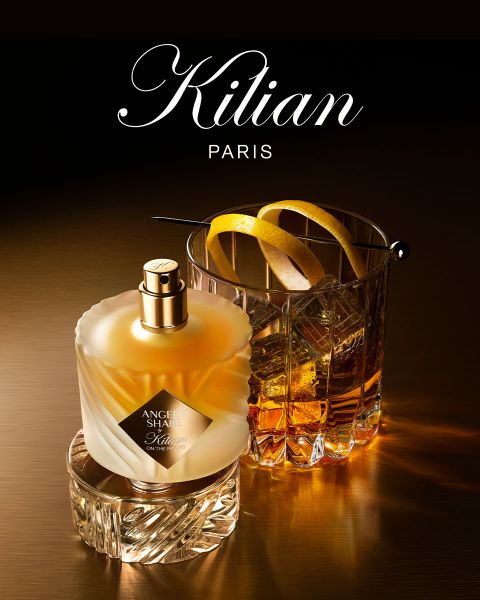 Ein Flakon des Parfums Kilian Paris Angels' Share steht neben einem Glas Whiskey mit Eis und einer Garnitur, beides auf einer reflektierenden braunen Fläche mit sanfter Beleuchtung. Oben ist das Kilian Paris Logo zu sehen.