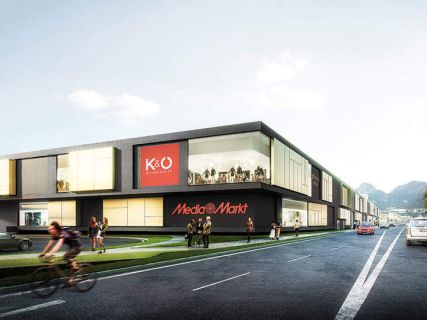 Modernes Geschäftsgebäude mit Media Markt- und K&Ö-Schildern, Menschen, die auf dem Bürgersteig gehen, und ein Radfahrer, der auf der Straße vorbeifährt.