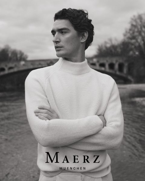 Ein Mann in einem weißen Strickpullover steht mit verschränkten Armen an einem Fluss, mit einer gewölbten Steinbrücke und kahlen Bäumen im Hintergrund. Der Text "MAERZ MUENCHEN" erscheint am unteren Rand des Schwarz-Weiß-Bildes.