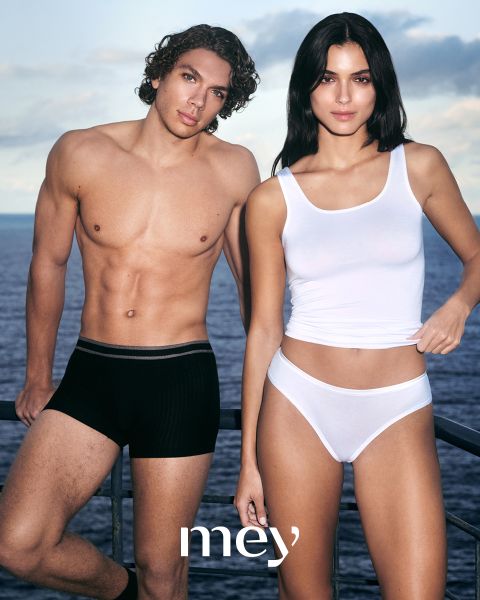 Ein Mann in schwarzen Boxershorts und eine Frau in einem weißen Tanktop und Unterwäsche stehen nebeneinander im Freien an einem Geländer, mit dem Meer und dem bewölkten Himmel im Hintergrund. Das Wort "mey" steht am unteren Rand.