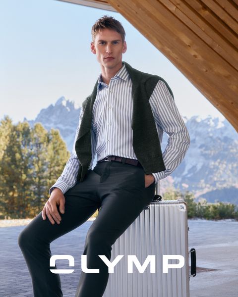 Ein Mann in einem gestreiften Hemd und einer dunklen Hose sitzt auf einem silbernen Koffer im Freien und hat sich einen Pullover über die Schultern gelegt. Im Hintergrund sind verschneite Berge und Bäume zu sehen. Am unteren Rand steht das Wort "OLYMP".