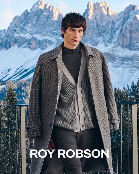 Ein Mann steht im Freien vor verschneiten Bergen und trägt einen grauen Hahnentrittmantel, einen grauen Pullover und eine graue Hose. Der Text "ROY ROBSON" erscheint am unteren Rand des Bildes.