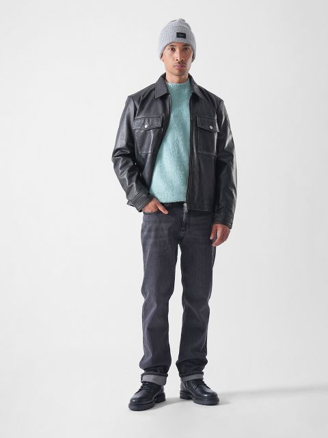 Ein Mann steht vor einem schlichten weißen Hintergrund, trägt eine graue Mütze, eine schwarze Lederjacke, einen hellblauen Pullover, dunkle Jeans und schwarze Schuhe und hat eine Hand in der Tasche.