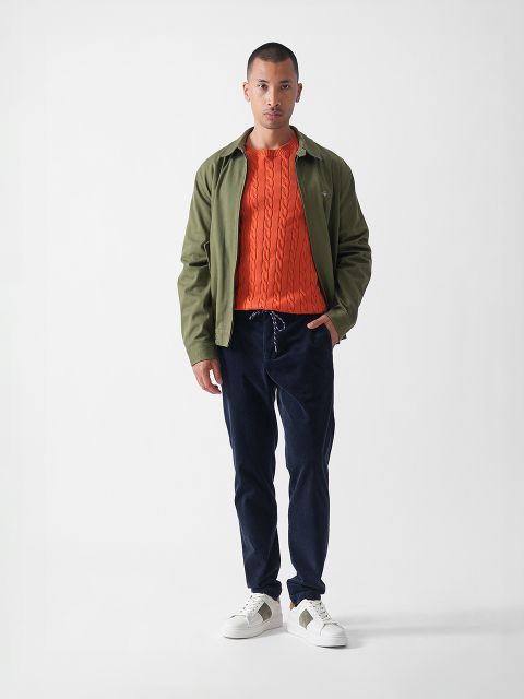 Ein Mann steht vor einem schlichten weißen Hintergrund und trägt eine olivgrüne Jacke über einem orangefarbenen Zopfstrickpullover, eine marineblaue Hose und weiße Turnschuhe mit grünen Akzenten.