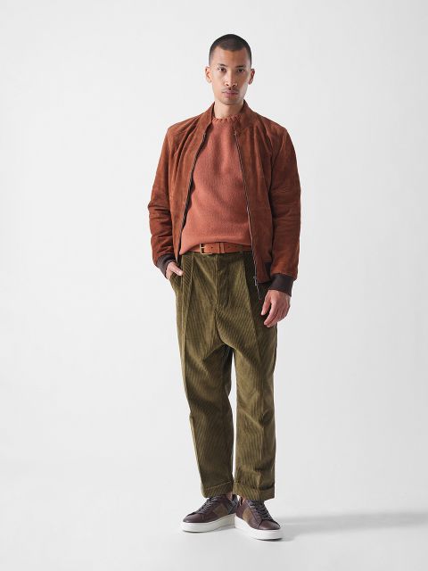Ein Mann steht vor einem schlichten weißen Hintergrund und trägt eine rostfarbene Wildlederjacke, einen dazu passenden Pullover, eine olivgrüne Kordhose, einen braunen Gürtel und braune Turnschuhe. Er schaut mit neutralem Blick direkt in die Kamera.
