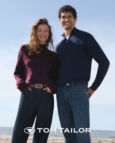 Eine lächelnde Frau in einem kastanienbraunen Pullover und ein Mann in einem marineblauen Pullover stehen Seite an Seite an einem Strand. Beide tragen Jeans. Der Himmel ist klar und blau. Das Logo "Tom Tailor" erscheint am unteren Rand.