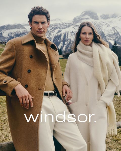 Ein Mann in einem braunen Mantel und eine Frau in einem cremefarbenen Mantel und Schal stehen im Freien mit Bergen im Hintergrund. Das Wort "windsor." ist am unteren Rand des Bildes zu lesen.