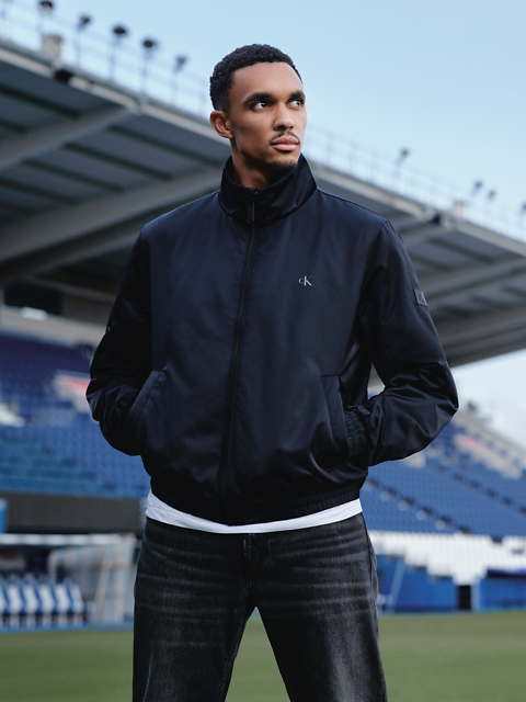 Ein Mann in schwarzer Jacke und Jeans steht in einem Stadion und schaut mit den Händen in den Taschen zur Seite. Im Hintergrund sind Stadionsitze und ein großes Gebäude zu sehen.