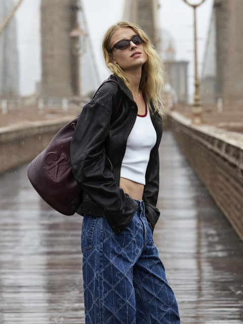 Eine Frau mit Sonnenbrille, schwarzer Jacke, weißem Crop-Top und weiten blauen Jeans steht auf einer verregneten Brücke und trägt eine große kastanienbraune Tasche über der Schulter. Die Brücke und die Lampen sind im Hintergrund unscharf.