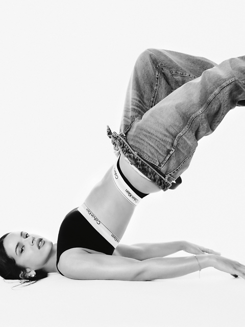 Schwarz-Weiß-Foto einer Frau in einem Calvin Klein Sport-BH und Jeans, die auf dem Rücken liegt, die Beine über den Kopf angewinkelt, die Arme auf dem Boden ausgestreckt, den Blick in die Kamera gerichtet.