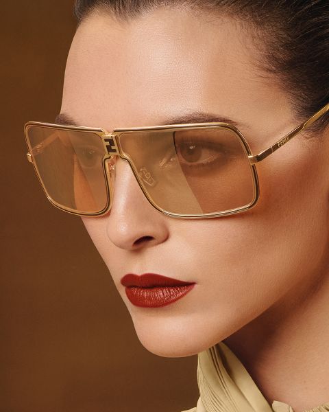 Nahaufnahme einer Person, die eine große, quadratische, goldgerahmte Sonnenbrille mit brauner Tönung trägt. Sie hat glatte Haut, zurückgekämmtes Haar, trägt kräftigen roten Lippenstift und ein helles Oberteil vor einem braunen Hintergrund.