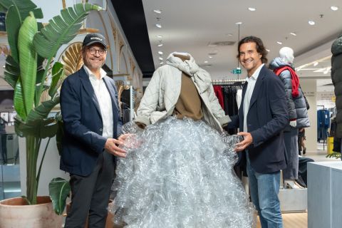 Zwei Männer in Blazern stehen neben einer Schaufensterpuppe, die eine Jacke und einen Rock aus durchsichtigen Plastikflaschen trägt, in einem modernen Bekleidungsgeschäft.