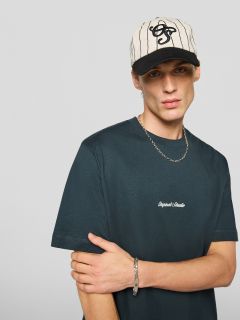 Ein junger Mann, der ein schwarzes T-Shirt mit der Aufschrift "Original Studio", eine silberne Halskette, ein silbernes Armband und eine creme-schwarz gestreifte Baseballkappe trägt, steht vor einem schlichten hellgrauen Hintergrund.