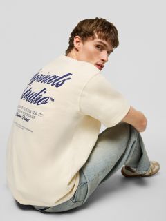 Ein junger Mann mit kurzen braunen Haaren, der ein cremefarbenes T-Shirt mit marineblauem Aufdruck auf dem Rücken, hellblaue Jeans und beigefarbene Turnschuhe trägt, sitzt auf dem Boden und schaut über die Schulter in die Kamera.