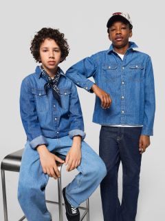 Zwei Jungen in Jeans und Jeanshemden posieren zusammen. Der eine sitzt auf einem Hocker, hat die Hände in den Schoß gelegt und einen Schal um den Hals, während der andere mit dem Arm auf dem Hocker steht und eine Mütze trägt.