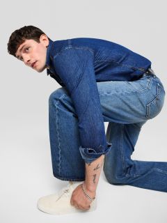 Ein junger Mann in Jeansjacke und Jeans kniet nieder und schaut zur Seite. Er trägt weiße Turnschuhe und hat eine sichtbare Tätowierung auf seinem Unterarm. Der Hintergrund ist einfarbig hellgrau.
