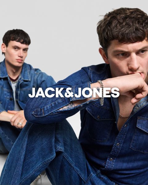 Zwei Männer in Jeans-Outfits sitzen vor einem schlichten Hintergrund. Der vordere Mann hält sich die Hand auf den Mund, während der andere hinter ihm sitzt. In der Mitte steht der Text "JACK & JONES".