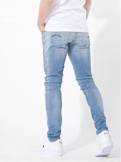 Eine Person, die ein weißes T-Shirt, hellblaue Skinny-Jeans und weiße Turnschuhe trägt, steht mit den Händen in den Hosentaschen, von der Kamera abgewandt, vor einem schlichten weißen Hintergrund.