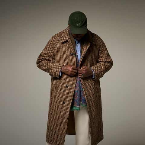 Eine Person mit grüner Baseballmütze, braunem Hahnentritt-Mantel, blauem Hemd, gemustertem Schal und heller Hose steht vor einem schlichten Hintergrund und schaut nach unten, während sie den Mantel zuknöpft.