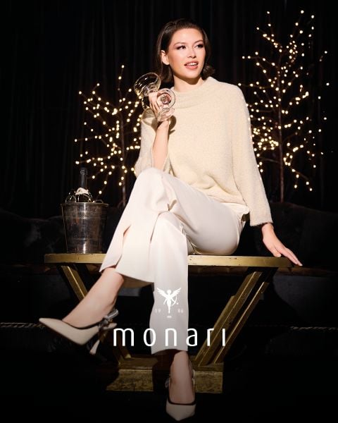 Eine Frau in einem cremefarbenen Pullover und einer weißen Hose sitzt auf einem goldenen Tisch und hält ein Weinglas in der Hand. Hinter ihr sind funkelnde Lichter, ein Eiskübel mit einer Flasche und das Logo "monari 1986" am Boden zu sehen.