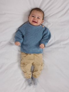 Ein lächelndes Baby liegt auf einer weißen Decke und trägt einen blauen Strickpullover, eine braune Cordhose mit Taschen und graue Socken. Das Baby sieht glücklich und entspannt aus und hat die Arme an den Ellbogen leicht angewinkelt.