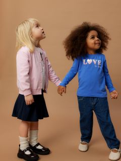 Zwei kleine Kinder stehen Händchen haltend vor einem einfarbig braunen Hintergrund. Ein Kind trägt eine rosafarbene Jacke und einen Rock, das andere eine blaue Jeans und ein blaues Hemd mit der Aufschrift "LIEBE IST KIND". Beide blicken neugierig nach oben.