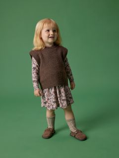 Ein kleines Kind mit blondem Haar steht lächelnd vor einem grünen Hintergrund und trägt einen braunen ärmellosen Pullover über einem geblümten Kleid, kniehohe Socken und braune Schuhe.