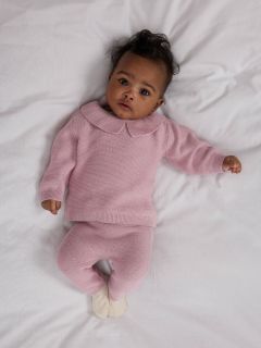 Ein Baby liegt auf einer weißen Decke und trägt einen hellrosa Strickpulli mit Petermännchenkragen und eine passende Hose sowie cremefarbene Socken.