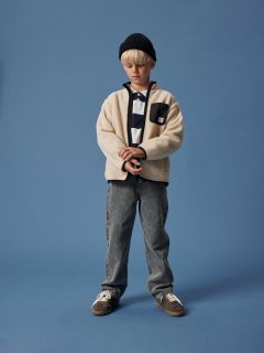 Ein Junge mit blondem Haar trägt eine schwarze Mütze, eine cremefarbene Fleecejacke über einem gestreiften Hemd, graue Jeans und Turnschuhe. Er steht vor einem einfarbig blauen Hintergrund und blickt mit gefalteten Händen nach unten.