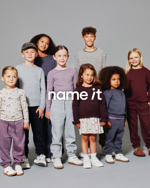 Eine Gruppe von acht verschiedenen Kindern steht vor einem grauen Hintergrund zusammen, alle tragen lässige, aufeinander abgestimmte Outfits in neutralen und Pastellfarben. Der Text "name it" erscheint in der Mitte des Bildes.