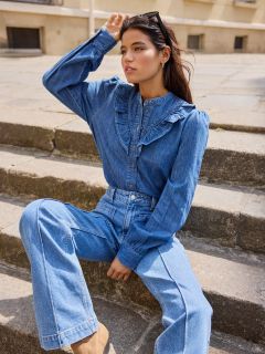 Frau, die auf einer Steintreppe im Freien sitzt und eine langärmelige Jeansbluse mit Rüschendetails und eine passende Jeans mit hoher Taille trägt. Sie hat dunkles Haar, eine Sonnenbrille auf dem Kopf und blickt nachdenklich in die Ferne.