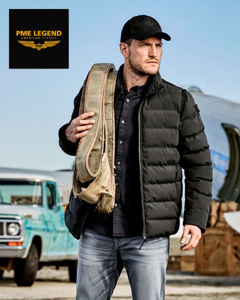 Ein Mann in schwarzer Pufferjacke, Mütze und Jeans trägt einen aufgerollten Segeltuchriemen über der Schulter und steht im Freien in der Nähe eines alten blauen Lastwagens und eines Flugzeugs. Das Logo von PME Legend erscheint in der oberen linken Ecke.