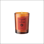 Eine orangefarbene Glaskerze mit der Aufschrift "Acqua di Parma Mosto" wird angezündet, wobei eine kleine Flamme an der Spitze sichtbar ist. Die Kerze steht vor einem schlichten weißen Hintergrund.